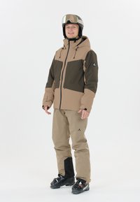 Giacca da sci beige e verde scuro con cappuccio e molte tasche, abbinata a pantaloni da sci beige con un accento nero e scarponi da sci.