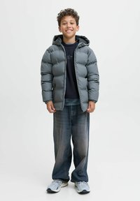 Grå puffjacka med huva, bärs över en mörk tröja, tillsammans med lösa blå jeans och grå sneakers. Leende barn poserar mot en vit bakgrund.