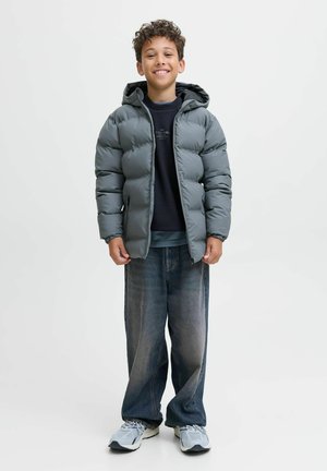 Doudoune grise avec capuche, portée sur une chemise sombre, associée à un jean bleu ample et des baskets grises. Enfant souriant prenant la pose devant un fond blanc.