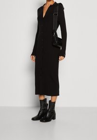 Abito a maniche lunghe in maglia nera con scollatura a V e bottoni frontali. Indossato con stivali neri alla caviglia e una piccola borsa a tracolla nera.