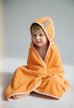 Kleinkind mit nassen Haaren, eingewickelt in ein orangefarbenes Kapuzenhandtuch mit einem Teddybär-Aufnäher, sitzend auf einer weißen Oberfläche.