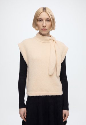 Femme blonde avec une coiffure courte au carré porte un pull sans manches crème avec un détail noué par-dessus un haut noir à manches longues et une jupe, face à l'avant.