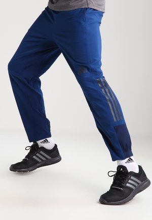 Niebieskie spodnie sportowe z szarymi paskami Adidas po bokach, elastycznym pasem i zwężanym krojem. Noszone z czarnymi butami sportowymi.