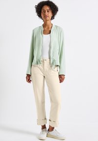Chaqueta bomber verde claro con cierre de cremallera, dobladillo con cordón y mangas largas, combinada con pantalones crema y zapatillas blancas.