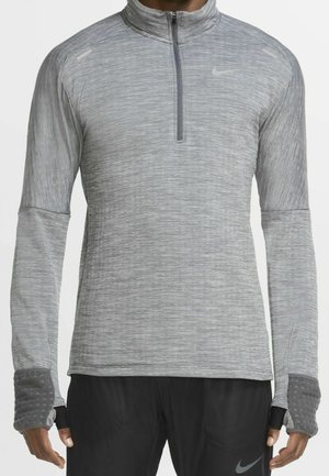 Person iført en lysegrå langærmet trøje med halv lynlås og tommelfingerhuller samt sorte bukser med et lille Nike-logo.