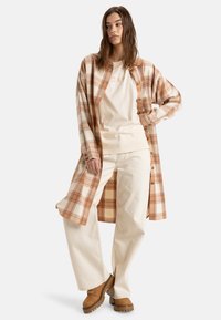 Plaid button-up shirt in cream and orange tones, layered over a beige t-shirt, paired with light-colored wide-leg pants and brown shoes.