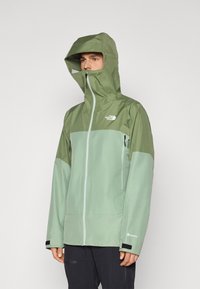 Veste GORE-TEX de couleur vert clair avec un panneau supérieur vert foncé, capuche ajustable, poches zippées et poignets en velcro. Tissu lisse et résistant à l'eau.