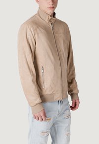 Giacca in pelle marrone chiaro con colletto alto, zip frontale e polsini a costine. Presenta due tasche con zip e una texture liscia simile alla suede.