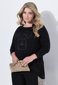 Schwarzes Langarmoberteil mit einem Strass-Floralmuster und dem Wort "chic". Das Model hält eine strukturierte goldene Clutch mit einer Schleifendetail.