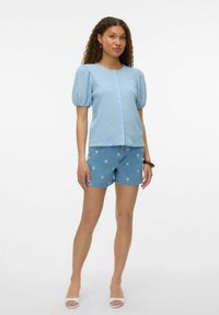 Blusa azzurro chiaro con texture e maniche a sbuffo corte; bottoni frontali. Abbinata a shorts di denim azzurro chiaro con ricami floreali. Sandali con tacco sottile.