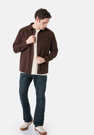 Mężczyzna zapinający ciemnobrązową koszulę na jasnobeżowy t-shirt, ubrany w ciemne dżinsy i białe trampki, stoi na jednolitym jasnym tle.
