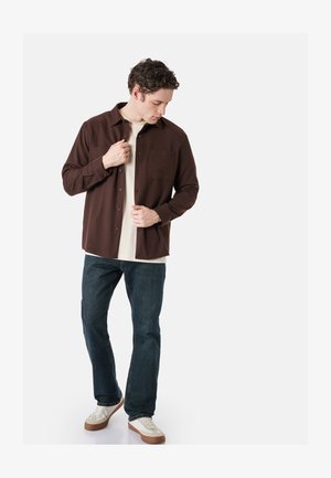 Mann knöpft ein dunkelbraunes Hemd über einem hellbeigen T-Shirt zu, trägt dunkle Jeans und weiße Sneaker, steht vor schlichtem hellem Hintergrund.