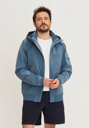 Man met een blauw gewatteerd jack met capuchon, wit T-shirt en zwarte korte broek, staand met gevouwen handen tegen een effen achtergrond.