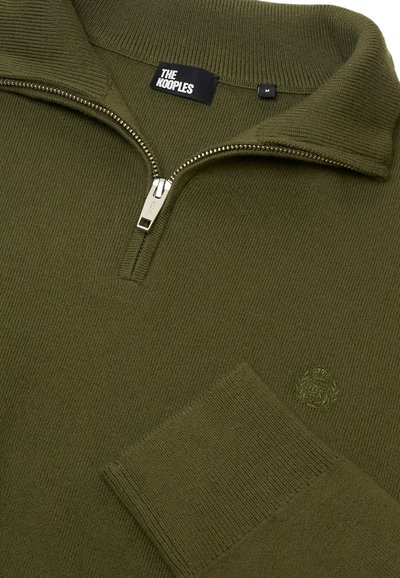 Olivgrüner Reißverschluss-Pullover mit gerippter Textur, der einen hohen Kragen und ein gesticktes Logo auf der Brust hat. Silberner Reißverschluss mit markiertem Zipper.