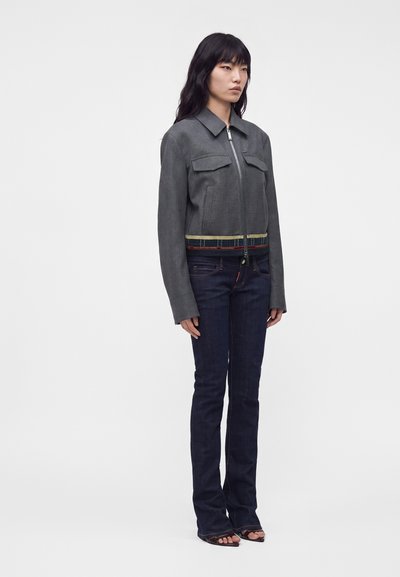 Femme debout portant une veste grise zippée avec des poches avant, un jean bleu foncé, et des sandales noires à bout ouvert sur un fond uni.