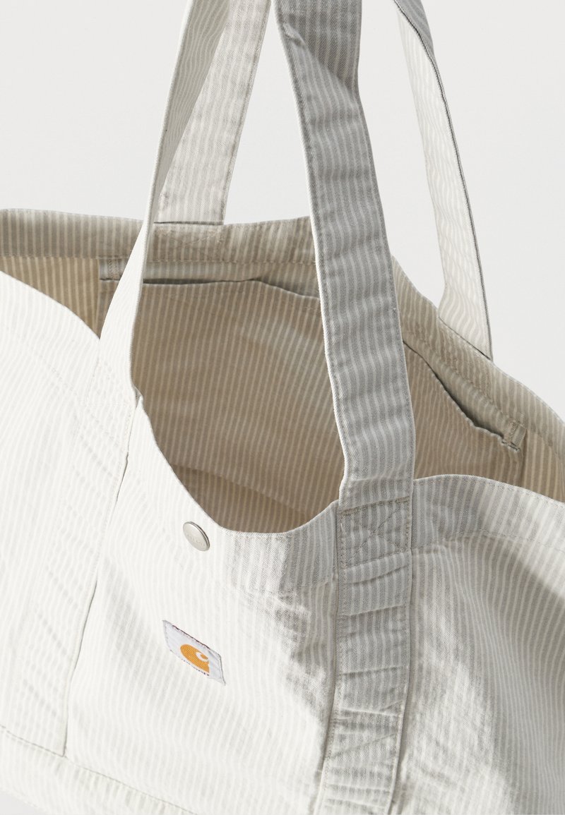 Witte en beige gestreepte stoffen draagtas met dubbele schouderbanden en een klein merklabel aan de voorkant, gezien van bovenaf geopend.