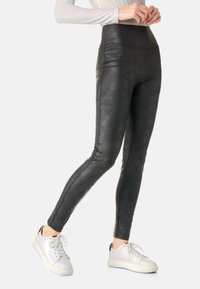 Schwarze Fake-Leder-Leggings mit hoher Taille, glatter Textur, eng anliegendem Design und Seitennahtdetails, kombiniert mit weißen Sneakern.