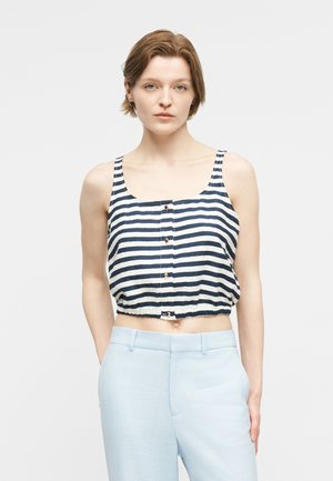 Žena v bezrukávovom námornícko modro-bielom pruhovanom krátkom topu s gombíkmi a svetlo modrých vysokých nohaviciach stojaca na jednoduchom pozadí.