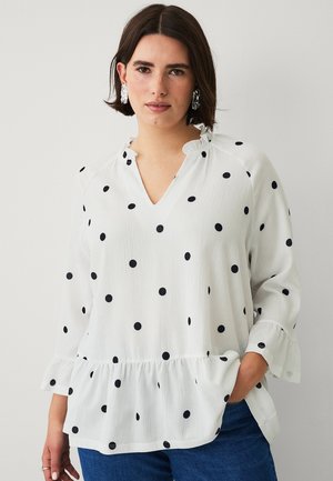 Kvinde iført hvid bluse med sorte prikker, lange ærmer, flæsekrave og -kant, kombineret med blå jeans, stående mod en ensfarvet baggrund.