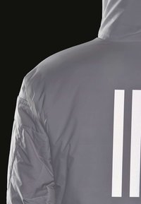 Veste grise à capuche avec deux bandes réfléchissantes blanches verticales dans le dos, présentée sur un fond noir.