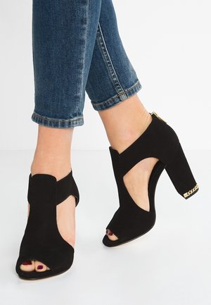 Füße in schwarzen Wildleder-Peeptoe-High-Heels mit goldener Kettendetail am Absatz, kombiniert mit hochgekrempelten blauen Jeans.