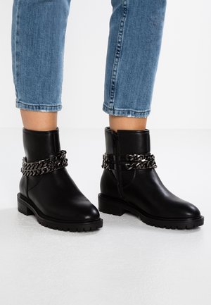 Czarne skórzane botki sięgające kostki z grubą podeszwą i dekoracyjnym srebrnym łańcuszkiem, noszone z krótkimi niebieskimi dżinsami.