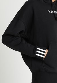 Musta huppari, joka on valmistettu puuvillasekoitteesta, jossa on kengurutasku, tekstuuriset hihansuut, joissa on kolme valkoista raitaa, sekä logo rinnassa.