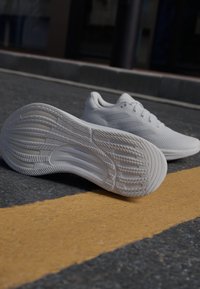 Zapatillas deportivas blancas con parte superior de malla texturizada y suela de goma. Presenta una suela curvada y un dibujo en la banda de rodadura para mayor tracción.