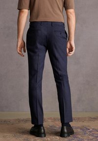 Férfi áll egy mintás szőnyegen, sötétkék, slim-fit nadrágban, barna, rövid ujjú ingben, fekete zokniban és fekete bőr cipőben, háttal állva.