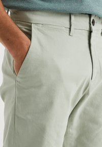 Chino din verde deschis cu o textură netedă, dotat cu buzunare laterale și închidere cu nasture; design minimalist cu o croială personalizată.