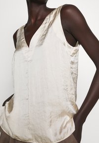 Top senza maniche beige chiaro con scollatura a V, realizzato in tessuto morbido. Il design presenta una vestibilità rilassata e una leggera lucentezza.