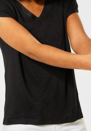 Persoon draagt een zwart T-shirt met korte mouwen en V-hals en strekt de armen over de borst uit, witte broek zichtbaar aan de onderkant.