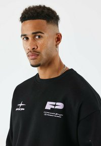 Jeune homme aux cheveux bouclés et piercings faciaux portant un sweat-shirt noir avec des logos et du texte blancs, regardant directement la caméra devant un fond blanc.