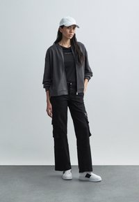 Szara bluza na zamek, czarna dopasowana koszulka, czarne spodnie cargo z bocznymi kieszeniami, białe trampki i biała czapka. Minimalistyczny design.