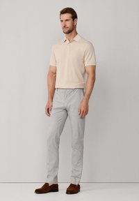 Uomo in piedi che indossa un polo beige a maniche corte, pantaloni grigi chiari e mocassini in suede marroni, su uno sfondo grigio chiaro uniforme.