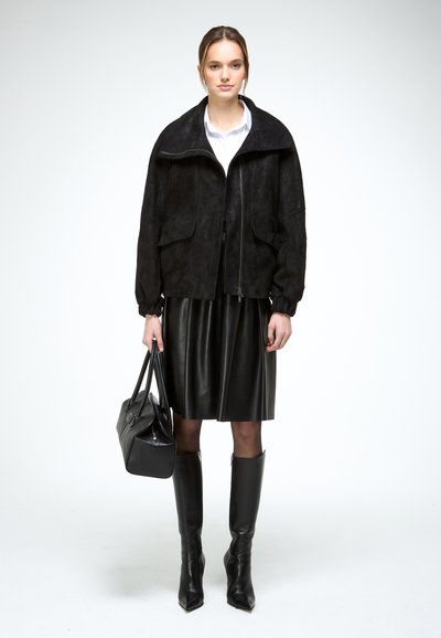 Femme portant une veste noire oversize, chemise blanche, jupe en cuir noire, bottes noires montant jusqu'aux genoux, tenant un sac à main noir, debout devant un fond uni.
