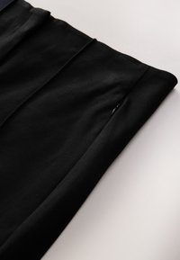 Black fabric pants featuring a smooth texture, pleated design, and a discreet zip pocket along the side seam.