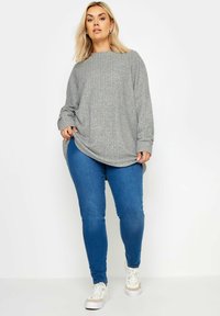 Übergroßer grauer Rippensweater mit langen Ärmeln, kombiniert mit schmalen blauen Jeans und weißen Sneakers. Die Textur ist weich und gemütlich.