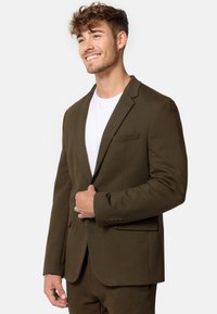 Blazer verde oliva con un petto singolo e rever a punta, dotato di una tasca sul petto e bottoni sui polsini, abbinato a una t-shirt bianca.