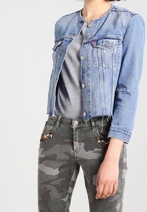Mujer con chaqueta vaquera corta de color azul claro sobre camiseta gris combinada con pantalones de camuflaje con detalles de mariposas bordadas.