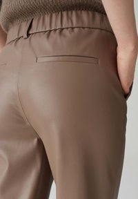 Bruine faux leren broek met een gladde textuur, voorzien van een geplooide tailleband en twee achterzakken. Het ontwerp is minimalistisch en getailleerd.