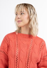 Korallfarbenes Strickpullover mit einem Kabeldesign und offenen Gewebedetails. Der Halsausschnitt ist weit und der Pullover hat ein lockeres, strukturiertes Aussehen.