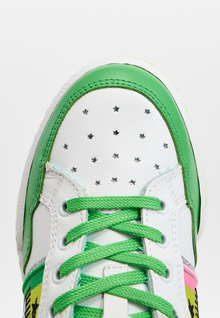 Zapatilla blanca con acentos verdes, perforaciones con diseño de estrellas, cordones verdes y una suela texturizada con paneles laterales vibrantes de colores múltiples.