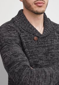 Dunkelgrauer gestrickter Pullover mit Schalkragen, strukturierten Mustern und einem braunen Knopfakzent am Hals.