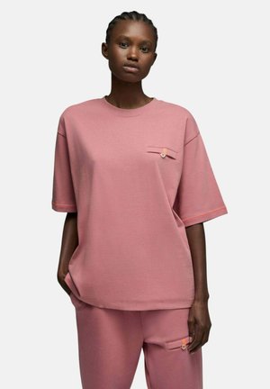 Femme aux cheveux tressés portant un haut rose ample à manches courtes et un pantalon assorti, chacun avec un détail de poche boutonnée, debout sur fond blanc.