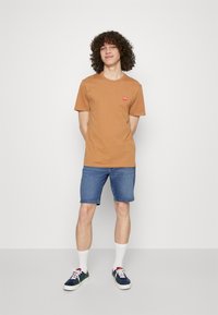Camiseta marrón de manga corta con un pequeño logo rojo, combinada con pantalones cortos de mezclilla azul, calcetines blancos y zapatillas multicolores.