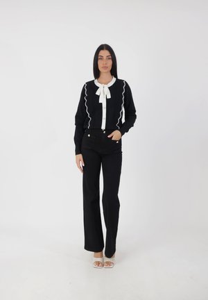 Zwarte cardigan met witte rand en knopen, strikdetail bij de kraag, gecombineerd met hoge zwarte broeken en doorschijnende hakken sandalen.