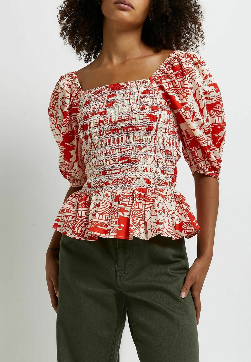 Femme portant un chemisier à manches bouffantes rouge et blanc à motifs, avec un corsage smocké et un ourlet à basque, associé à un pantalon vert foncé.