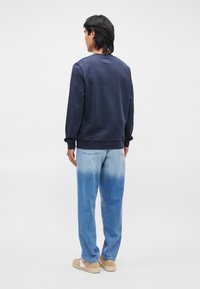 Sweatshirt azul escuro com punhos e bainha canelados, combinado com jeans em degradê azul claro. O modelo usa sapatos bege, visto de costas.