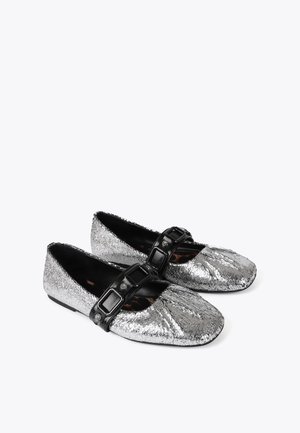 Silver ballet flats with a metallic texture, rounded toe, and a black buckle strap across the instep. Flat, flexible sole.